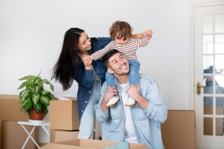 Portrait of happy family with little son having fun in new home after moving, cheerful parents and their cute toddler child bonding together, posing among cardboard boxes, celebrating relocation