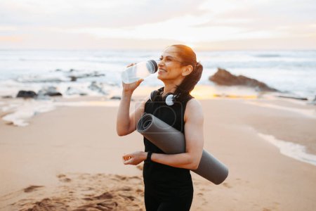 Photo for Smiling mature european woman in sportswear with mat, drinks bottle of water, enjoy workout in morning on sea beach, outdoor. Weight loss sport, fitness at sunrise and body care - Royalty Free Image