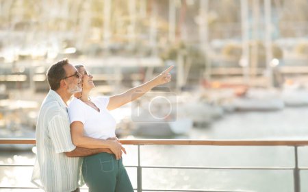 Téléchargez les photos : Vacances d'été. Joyeux retraités européens conjoints regardant les yachts de luxe à Marina, pointant du doigt à côté de l'espace vide debout à la jetée à l'extérieur. Aînés Publicité Offre de vacances - en image libre de droit
