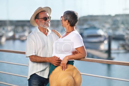 Téléchargez les photos : Vacances d'été Béatitude. Heureux couple de personnes âgées profite de l'été à Marina Dock en plein air, étreignant devant des yachts élégants, posant avec des chapeaux de paille et des lunettes de soleil. Joie des vacances à la mer - en image libre de droit