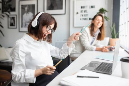 Photo for Joyful lady office manager in headphones enjoying music and singing while working on laptop in office, sitting with colleague at workplace, selective focus - Royalty Free Image