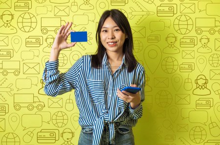 Foto de Sonriente linda mujer asiática joven con teléfono y tarjeta bancaria en sus manos sobre diversos iconos del mundo digital de fondo. Concepto de tecnologías modernas, transferencia de dinero, banca, pagos en línea, aplicación móvil - Imagen libre de derechos