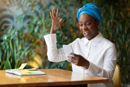 Photo for Thrilled happy beautiful young black woman entrepreneur sitting at table at cafe, looking at phone screen, gesturing, raising hand up, have great news, got grant, investment for business, copy space - Royalty Free Image