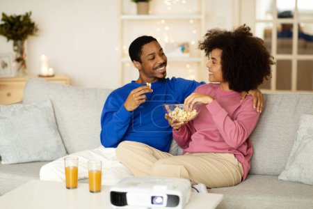 Photo for At Home Cinema. Happy Black Millennial Couple Watching Film Enjoying Movie Night Together, Eating Popcorn Sitting On Sofa Near Projector In Living Room Indoor. Modern Entertainment. Selective Focus - Royalty Free Image
