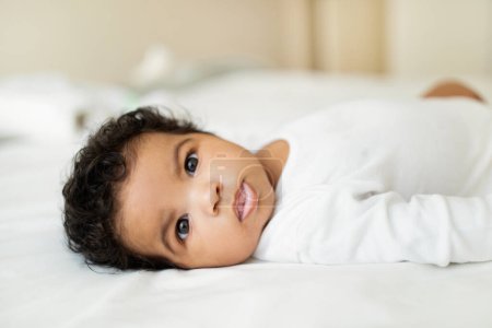 Photo for Happy cute sweet black little child lies on white bed in bedroom, kid rest and relax, close up. Newborn, childhood, baby care at home, ad and offer, health care and safe on mattress - Royalty Free Image