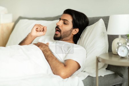 Photo for Unhappy sad middle eastern guy woke up, lying on bed, coughing, suffering from flu and cold in bedroom interior. Sick from virus, symptoms, health problems, medicine, ad and offer - Royalty Free Image