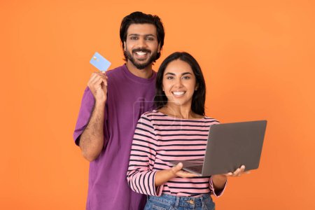 Photo for Virtual banking. Cheerful beautiful young hindu couple holding modern computer laptop and bank card, lovers paying for goods and services online, isolated on orange stuido background - Royalty Free Image
