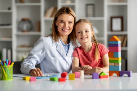 Photo for Portrait of happy little girl and child development specialist lady sitting at desk with colorful wooden bricks and smiling at camera, therapist woman and cute female kid playing and posing together - Royalty Free Image