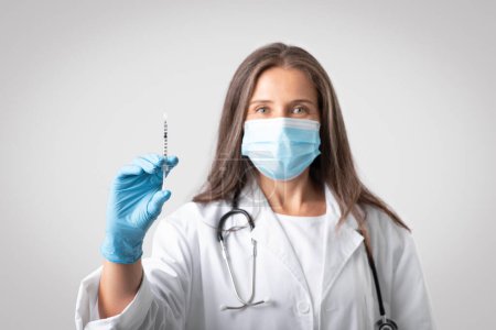 Photo for Senior woman therapist in white coat and protective mask showing syringe with vaccine, isolated on gray background, selective focus. Doctor doing vaccination, immunization for health care - Royalty Free Image