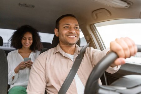 Téléchargez les photos : Conducteur de taxi arabe heureux naviguant voiture tenant roue, tandis que passager Lady SMS sur Smartphone assis sur le siège arrière dans l'automobile à l'intérieur. Offre de transport de véhicules Concept. Focus sélectif - en image libre de droit