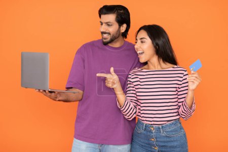 Photo for Happy beautiful loving young indian couple in casual holding modern pc laptop and blue plastic bank card, woman pointing at computer screen, lovers booking tickets online, orange background - Royalty Free Image