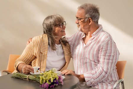 Téléchargez les photos : Vieil homme joyeux embrassant femme dans casual assis à la table, profiter du café ensemble dans le café en plein air. Amour, relations, temps libre et week-end, retraite et rendez-vous romantique à l'été - en image libre de droit