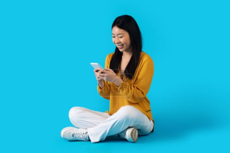 Photo for Entertainment and gadgets. Cheerful smiling attractive young asian woman using smartphone on blue studio background, checking newest mobile app, shopping, banking, studying online, copy space - Royalty Free Image