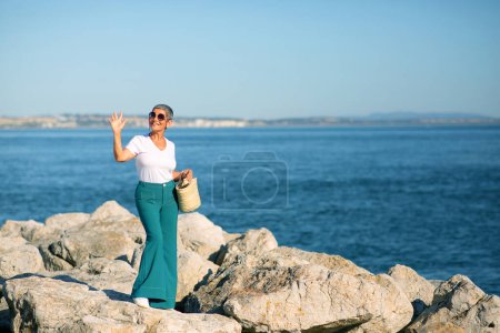 Téléchargez les photos : Bonjour, Summer. Happy European Mature Lady agitant la main à la caméra debout près de la côte de la mer en plein air. Femme Profitant de vacances et journée ensoleillée au bord de la mer. Concept de vacances de rêve. Longueur totale - en image libre de droit