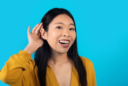 Photo for Beautiful cheerful young asian woman holding hand near ear, trying to listen to interesting news expressing communication concept and gossip, blue studio background, copy space, closeup - Royalty Free Image