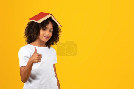 Photo for Smiling adolescent curly african american girl in white t-shirt with book on head show thumb up, isolated on yellow studio background. Approving, recommendation reading, study, knowledge, offer, ad - Royalty Free Image
