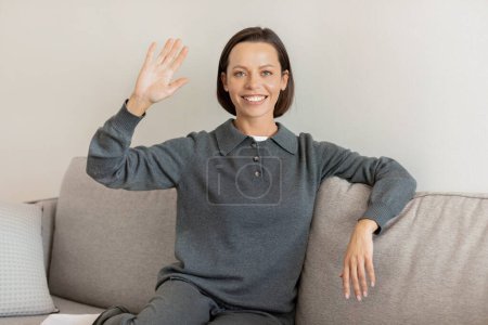 Photo for Positive pretty caucasian adult lady in domestic clothes sits on sofa, waving hand in living room interior. Video call, housewife resting at home, enjoy comfort alone, ad and offer - Royalty Free Image