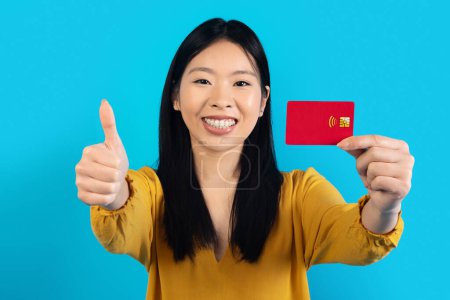Photo for Happy pretty millennial asian woman with braces and in casual showing red credit card and thumb up at camera, smiling, recommending contactless payment, easy fast banking, blue studio background - Royalty Free Image