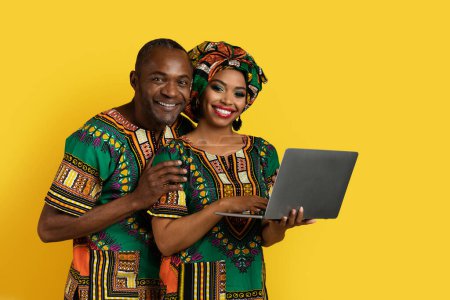 Photo for Happy beautiful black couple wearing national african costumes websurfing on laptop pc over yellow studio background, embracing and smiling at camera, have loving bond, copy space - Royalty Free Image