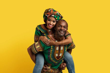 Photo for Ceerful young beautiful black woman piggybacking her man, loving cute couple wearing african costumes enjoying time together on yellow studio background, copy space. Love, affectionate concept - Royalty Free Image