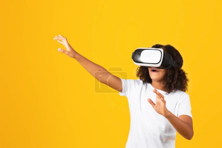 Photo for Cheerful shocked surprised adolescent curly black girl in white t-shirt and vr glasses touches empty space with hands, isolated on yellow studio background. Fun, entertainment device, emotions - Royalty Free Image