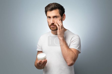 Photo for Grooming essentials. Middle aged man applying moisturising cream on face while standing on grey studio background. Handsome male using nourishing lotion for skin - Royalty Free Image