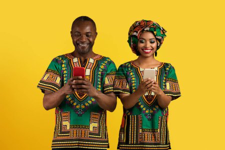 Photo for Gadget addiction and relationships concept. Happy black couple in national african costumes using smartphones on yellow studio background, scrolling, checking social media - Royalty Free Image