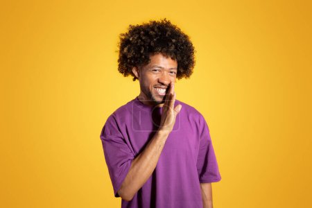 Photo for Positive handsome middle aged black curly man in purple t-shirt say secret gesture isolated on yellow background, studio. Gossip, ad and offer, human emotions and good news - Royalty Free Image