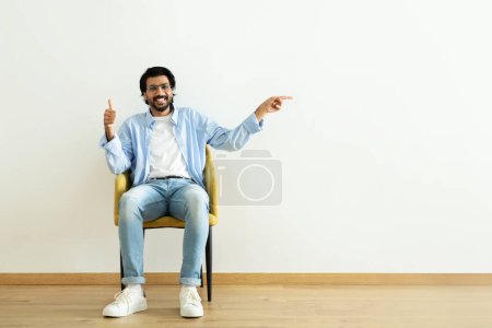 Foto de Positivo millennial Oriente Medio chico mostrar el pulgar hacia arriba, aprobando, señalando el dedo en el espacio vacío, sentarse en el sillón en la habitación en el fondo de la pared blanca. Recomendación, asesoramiento, anuncio y oferta - Imagen libre de derechos