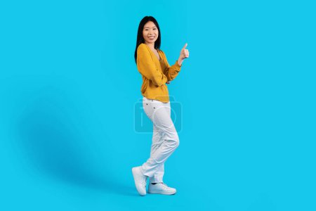 Foto de Alegre hermosa mujer coreana joven despreocupada con traje casual que muestra el pulgar hacia arriba y sonriendo a la cámara, recomendando un buen trato, aislado en el fondo del estudio azul, espacio de copia, longitud completa - Imagen libre de derechos