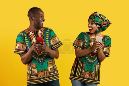 Photo for Surprised cheerful black middle aged man and young woman looking at each other and smiling over yellow studio background, couple wearing african costumes, using cell phones. copy space - Royalty Free Image