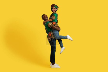 Photo for Loving cheerful handsome strong black man husband lifting up his pretty wife smiling young woman, couple wearing national african costumes, enjoying time together on yellow background, copy space - Royalty Free Image