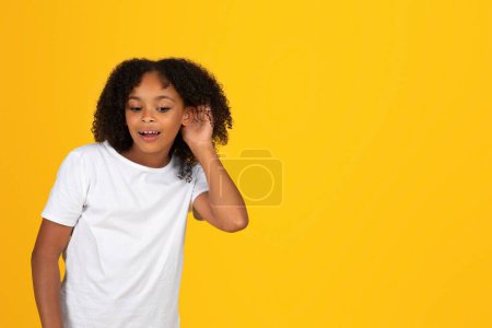 Téléchargez les photos : Souriant jeune fille bouclée intéressée en t-shirt blanc écoute secret, isolé sur fond de studio jaune. commérages, bonnes nouvelles, publicité et offre, connaissances scolaires, émotions des élèves - en image libre de droit