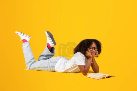 Photo for Positive smart teen black curly girl in white t-shirt, glasses reads book, lies on floor, isolated on yellow studio background. Study, knowledge, hobby, rest and relax, ad and offer - Royalty Free Image