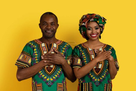 Photo for Cheerful smiling african american lovers middle aged man and young woman in traditional costumes holding hands on chest over heart, expressing love and affection, yellow studio background - Royalty Free Image