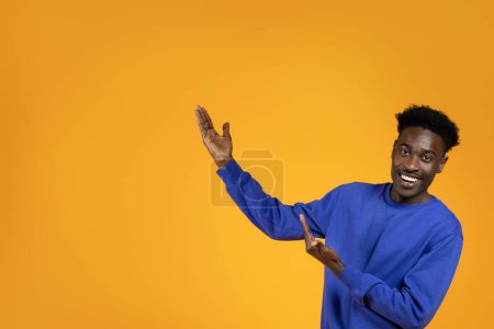 Photo for Attractive happy smiling millennial black man in stylish outfit showing copy space for text or advertisement on yellow studio background. African american guy recommending nice offer or deal - Royalty Free Image