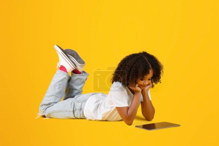 Photo for Smiling adolescent curly black girl in white t-shirt watch video lesson, plays online game on tablet, lies on floor, isolated on yellow studio background. App for study, device for video call - Royalty Free Image