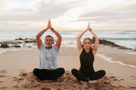 Photo for Concentrated mature european family in sportswear practice yoga, meditate, enjoy workout together in morning, sit on sea beach, outdoor. Peace, sports, life style and body care, asana and breath - Royalty Free Image
