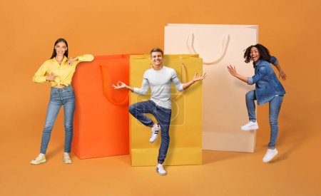 Photo for Multicultural friends emotional millennial two ladies and one guy gesturing next to huge shopping bags over colorful studio background, celebrating season sale, mockup, collage, full length - Royalty Free Image