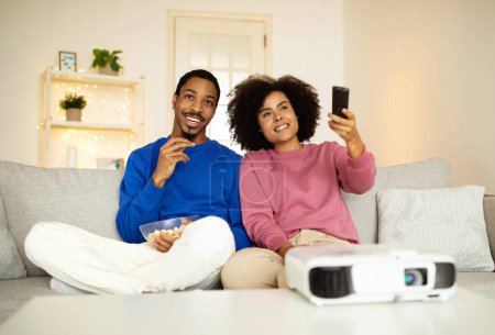 Foto de Alegre pareja joven afroamericana viendo la película juntos utilizando proyector, apuntando controlador remoto en casa, abrazando sentado en el sofá interior. Cónyuges relajante disfrutando de buena película en fin de semana - Imagen libre de derechos