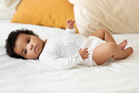 Photo for Serious black little child girl or boy in clothes lies on white comfort bed in bedroom. Health care, newborn, childhood, child care, parenthood at home, ad and offer, full length - Royalty Free Image
