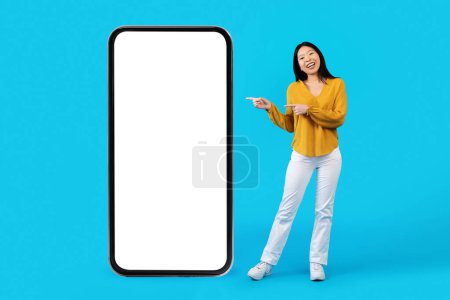 Photo for Cheery smiling young asian lady in casual pointing at huge phone with white empty screen, mockup of ad, isolated on blue studio background, copy space. Shopping mobile app, e-commerce - Royalty Free Image