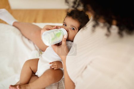 Photo for Black millennial mother feeds from bottle hungry little baby on white bed, enjoys love and comfort in bedroom. Kid food with vitamins, baby care, parenthood and family at home - Royalty Free Image