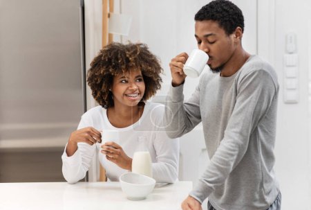 Photo for Time together, relationships, marriage, affectionate. Happy young black lovers wearing pajamas drinking coffee at kitchen, have conversation, smiling, copy space for advertisement - Royalty Free Image