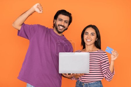 Photo for Cashback on bank card. Emotional young eastern man and woman in casual outfits posing with bank card and laptop on orange studio background, guy clenching fist, celebrating success - Royalty Free Image