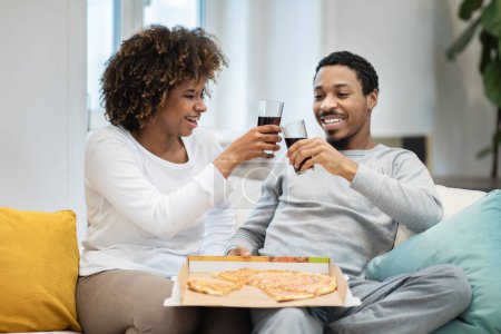 Téléchargez les photos : Cheerfy beaux jeunes amateurs noirs ont dîner à la maison. Heureux homme et femme afro-américain en tenue de maison assis sur le canapé, manger de la pizza, acclamer avec des lunettes, boire, célébrer bonne journée - en image libre de droit