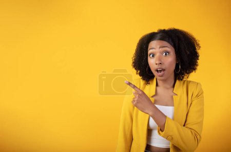 Foto de Sorprendido joven dama negra señalando con el dedo a un lado en el espacio libre aislado en el fondo del estudio amarillo, recomendando una gran venta, grandes noticias, espacio para el anuncio. Personas emociones y sorpresa - Imagen libre de derechos