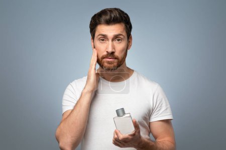 Photo for Grooming mastery. Handsome middle aged caucasian man applying perfume on beard after shaving, standing over grey studio background and looking at camera - Royalty Free Image