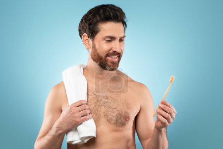 Photo for Handsome middle aged man with towel on shoulder holding toothbrush, enjoying oral hygiene, posing on blue background. Good morning, beauty care, daily procedures, caries control - Royalty Free Image
