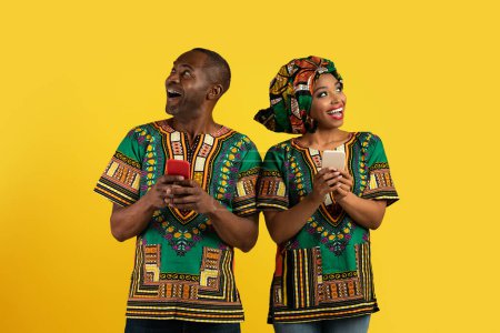 Photo for Excited black couple wearing traditional african costumes with smartphones in their hands looking aside at copy space for text or advertisement over yellow studio background - Royalty Free Image